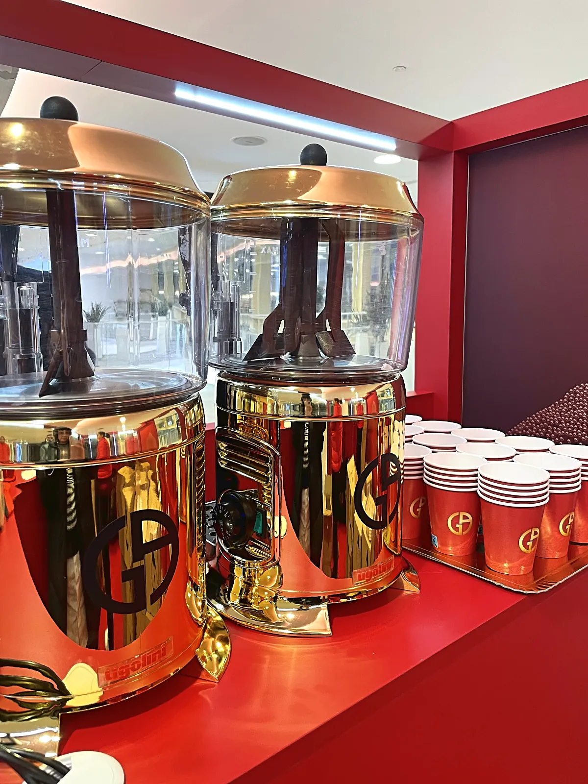 Gros plan sur deux chocolatières dorées au logo GA et pile de gobelets rouges